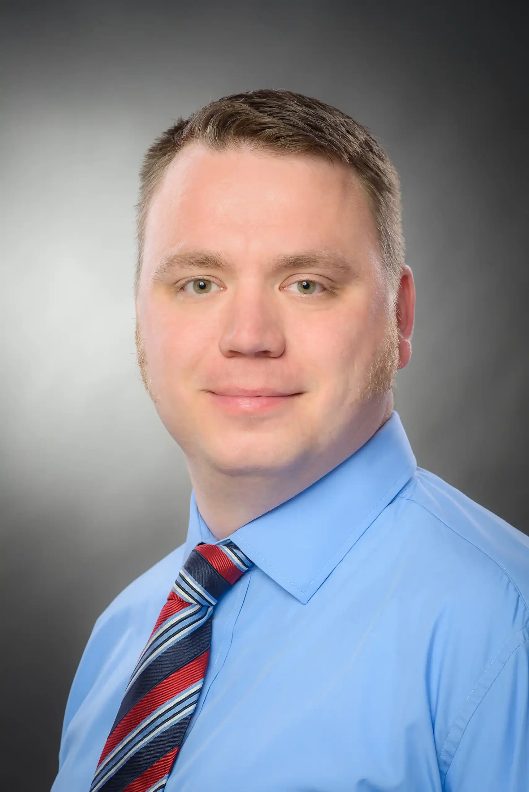 Bernd Kinny mit kurzen hellbraunen Haaren, blauem Hemd und gestreifter Krawatte posiert für ein professionelles Portraitfoto vor einem neutralen Hintergrund.