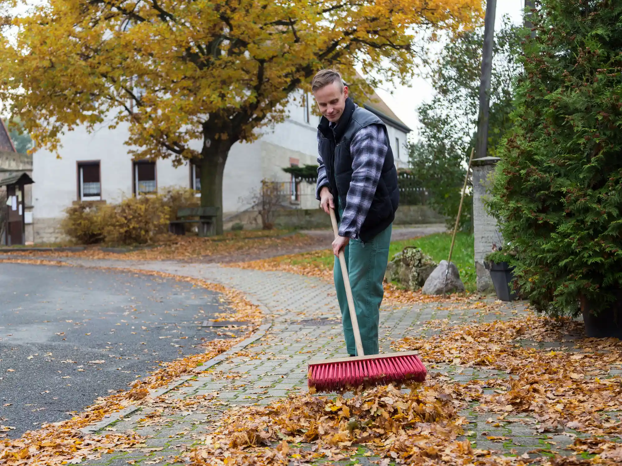 Eine Person harkt herabgefallene Herbstblätter auf einem gepflasterten Gehweg neben einer Straße, mit Bäumen und Gebäuden im Hintergrund.